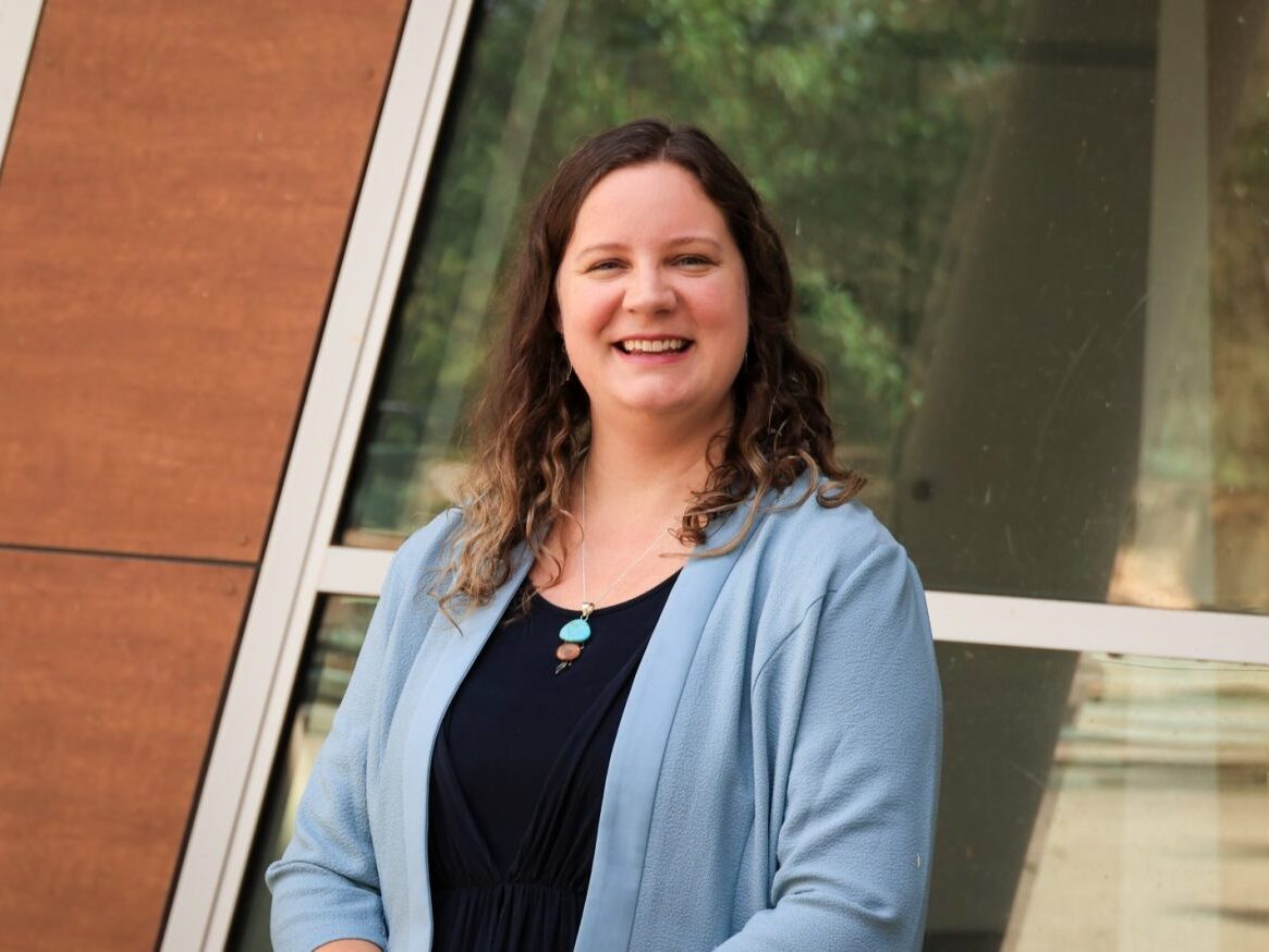 A photo of Laura, from the waist up. She is wearing a black top with a blue blazer over the top. She has brown, curly hair. It falls just past her shoulders. She is smiling at the camera. She is standing in front of a building.