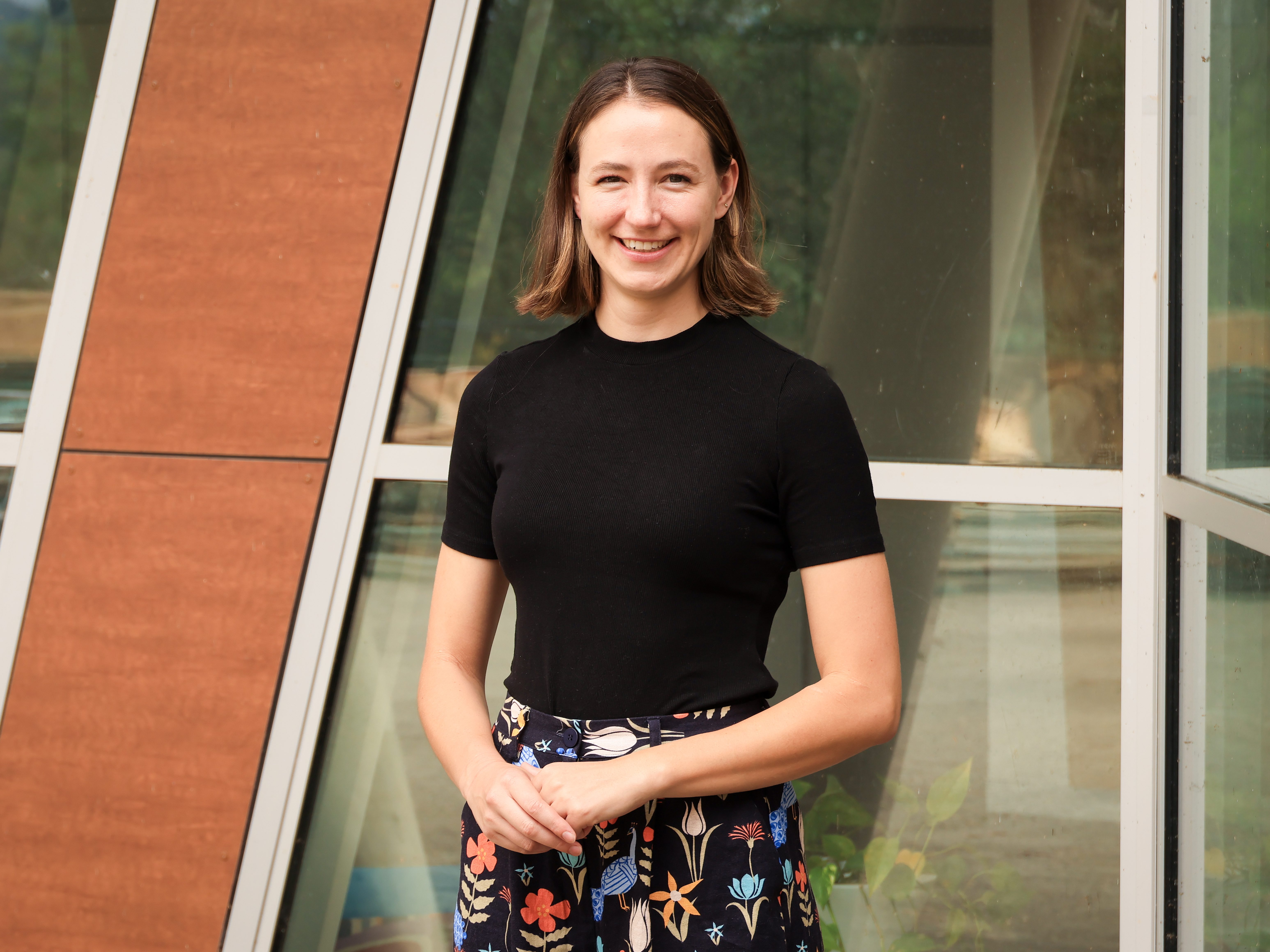 A photo of Molly from the waist up. She has brown, shoulder-length hair. She is wearing a black top and trousers with bright flowers on them. Her hands her held together in front of her. She is standing in front of a building.