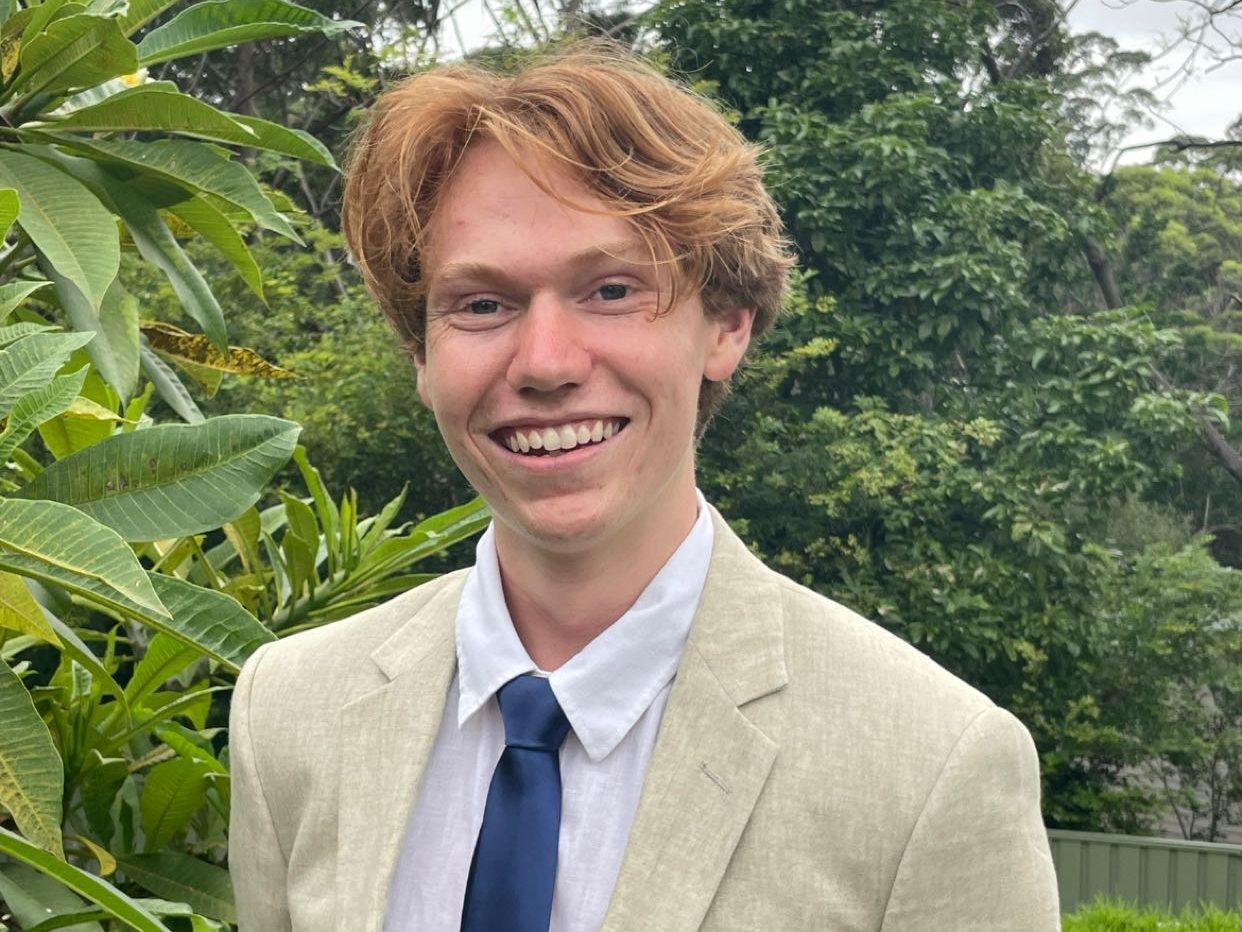A photo of Talon, from the chest up. He is wearing a cream blazer and a blue tie. He has red hair and blue eyes. He is smiling widely. The background is green trees. 