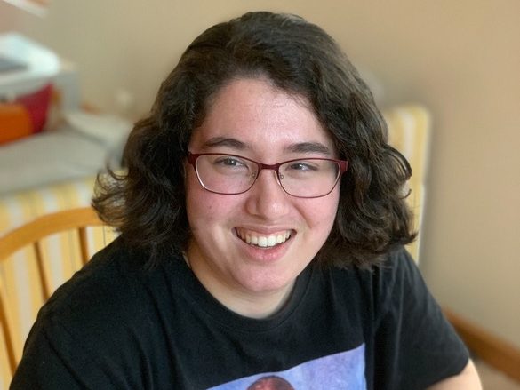 A portrait photo of Meg. She has dark, curly, shoulder-length hair. She is wearing glasses and a black t-shirt. She is smiling at the camera. 